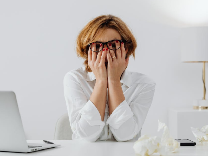 A stressed-out woman sitting at her desk in front of a laptop, and rubbing her eyes beneath her glasses.