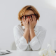 A stressed-out woman sitting at her desk in front of a laptop, and rubbing her eyes beneath her glasses.