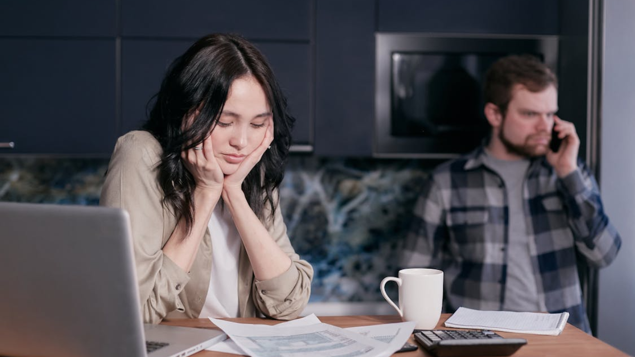 A tired woman looking at documents and a laptop on a kitchen table, while a man talks on the phone in the background.